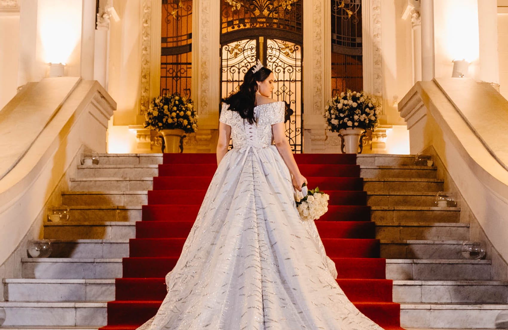 Noiva no alto da escadaria do salão, com a cauda do vestido estendida sobre o tapete vermelho, em casamento produzido pela Rito Eventos